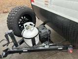 Detail of an RV rental trailer's hitch area, featuring an off-road spare tire, propane tank, and battery.