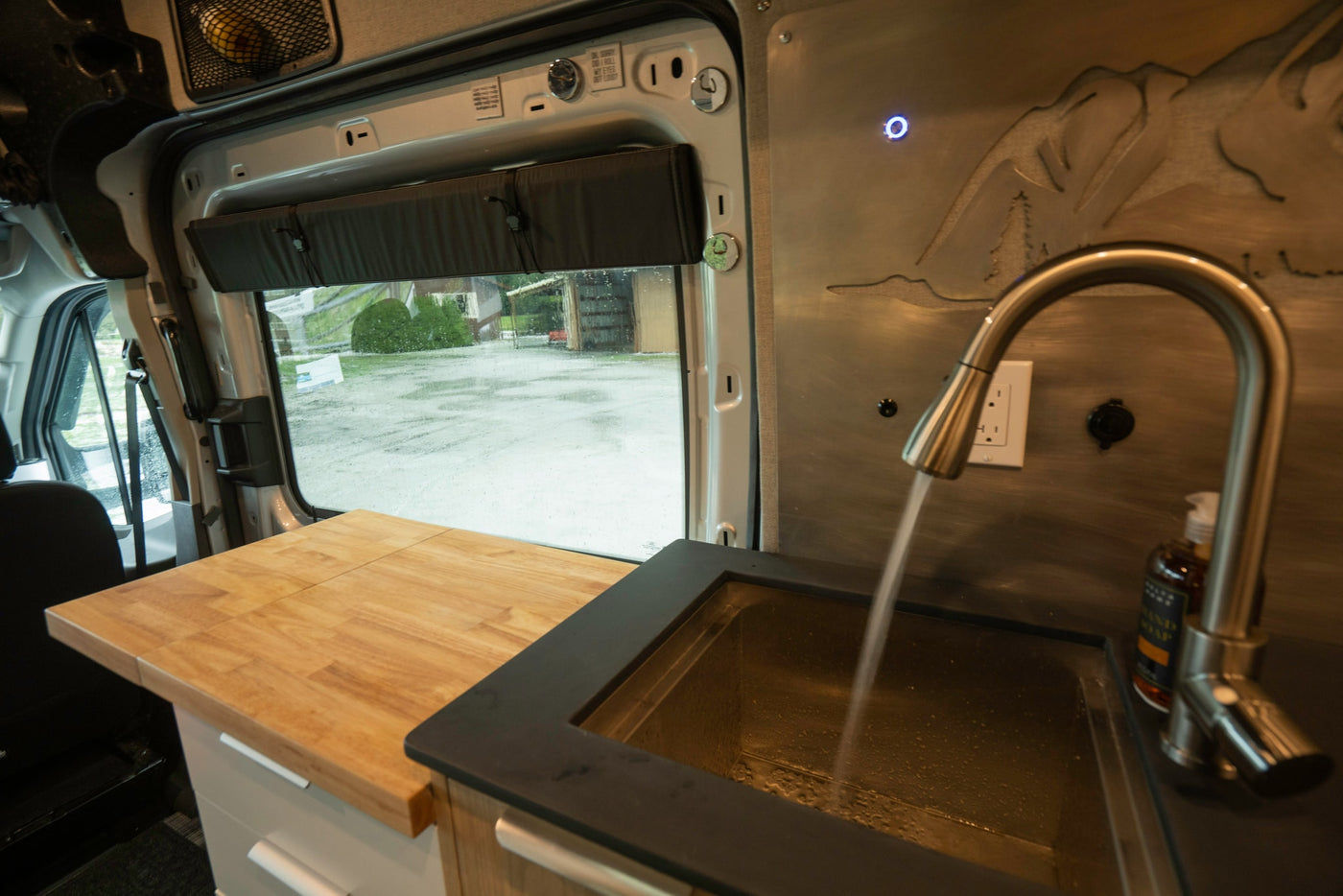 Inside an RV rental, a compact kitchen with a running water sink, wood counter, and window view of a rainy day. — photo 14