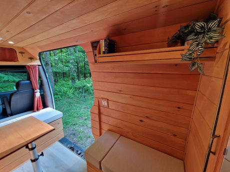 Wood-paneled RV rental interior, showing cozy seating, a bookshelf with plants, and a forest view from the open door.