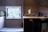 This RV rental interior features a compact kitchenette with a sink, fridge, and kettle, beside a cozy sleeping area and window.