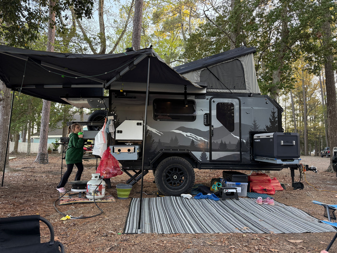 A modern RV rental travel trailer with a pop-up top and awning at a wooded campsite, featuring outdoor cooking gear.