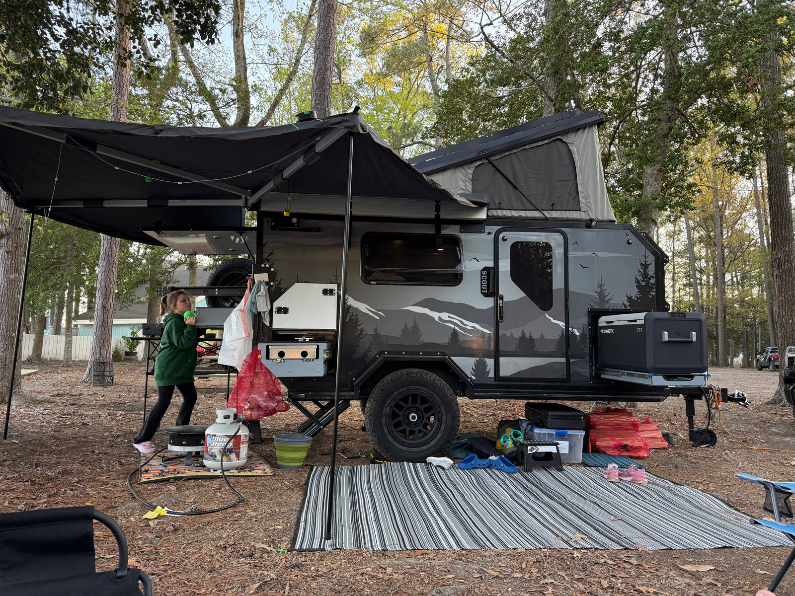 A modern RV rental travel trailer with a pop-up top and awning at a wooded campsite, featuring outdoor cooking gear.