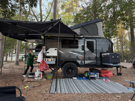 A modern RV rental travel trailer with a pop-up top and awning at a wooded campsite, featuring outdoor cooking gear.