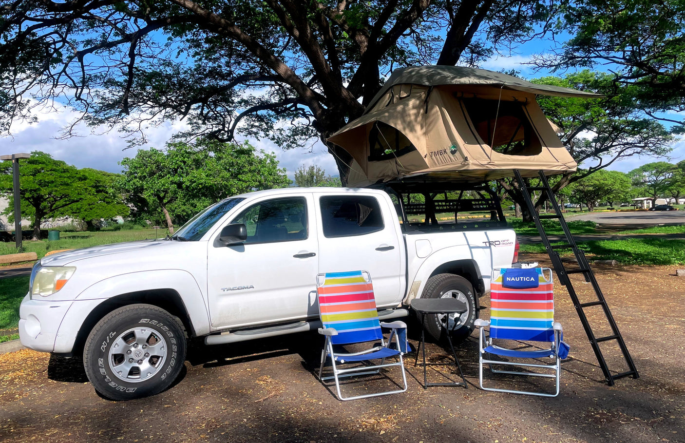 White pickup truck with rooftop tent, chairs, & table parked in a leafy area, showing a unique RV rental camping setup.