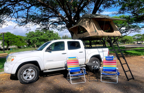 White pickup truck with rooftop tent, chairs, & table parked in a leafy area, showing a unique RV rental camping setup.