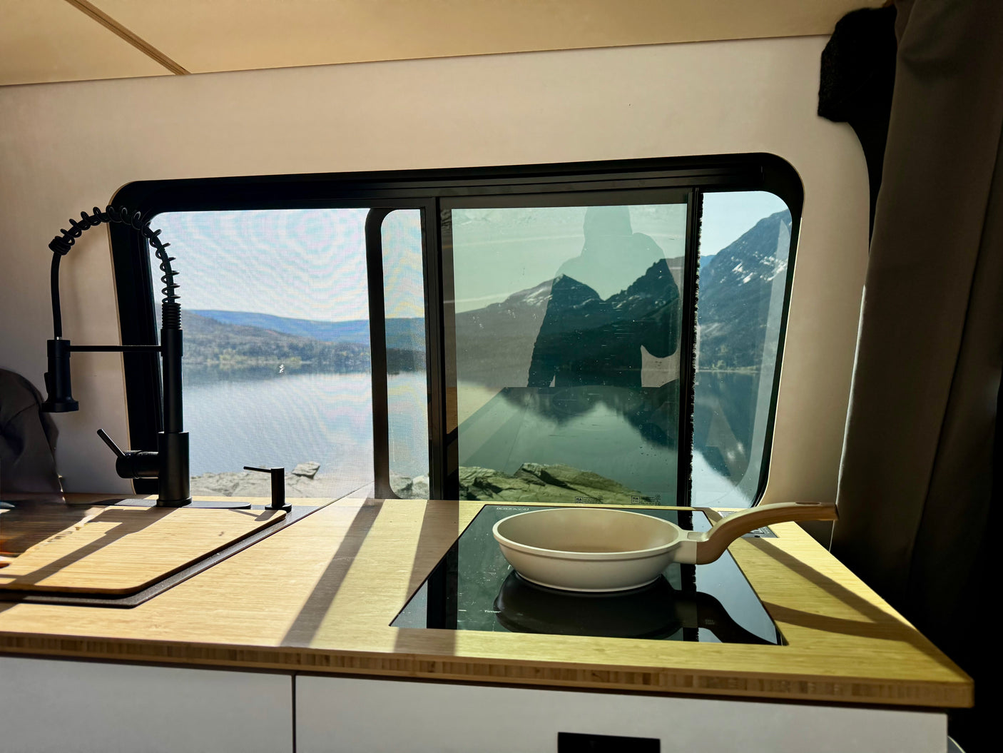 Modern rv rental kitchen with a large window revealing a serene lake and majestic mountains. Pan on induction cooktop.