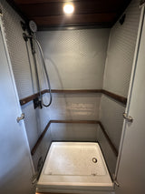 Compact RV rental shower stall with grey hexagon tiles, wood trim, and a white shower pan, featuring a black handheld shower.