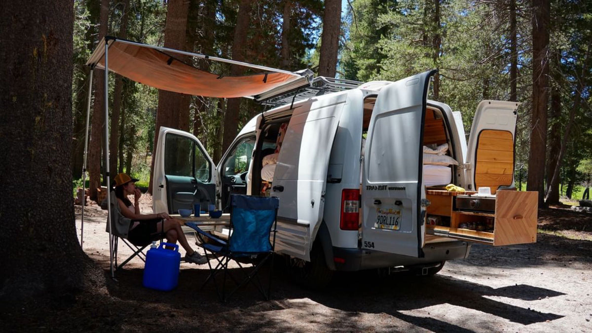 RV rental adventure: Camper van with awning & pull-out kitchen, a person relaxing at a forest campsite.