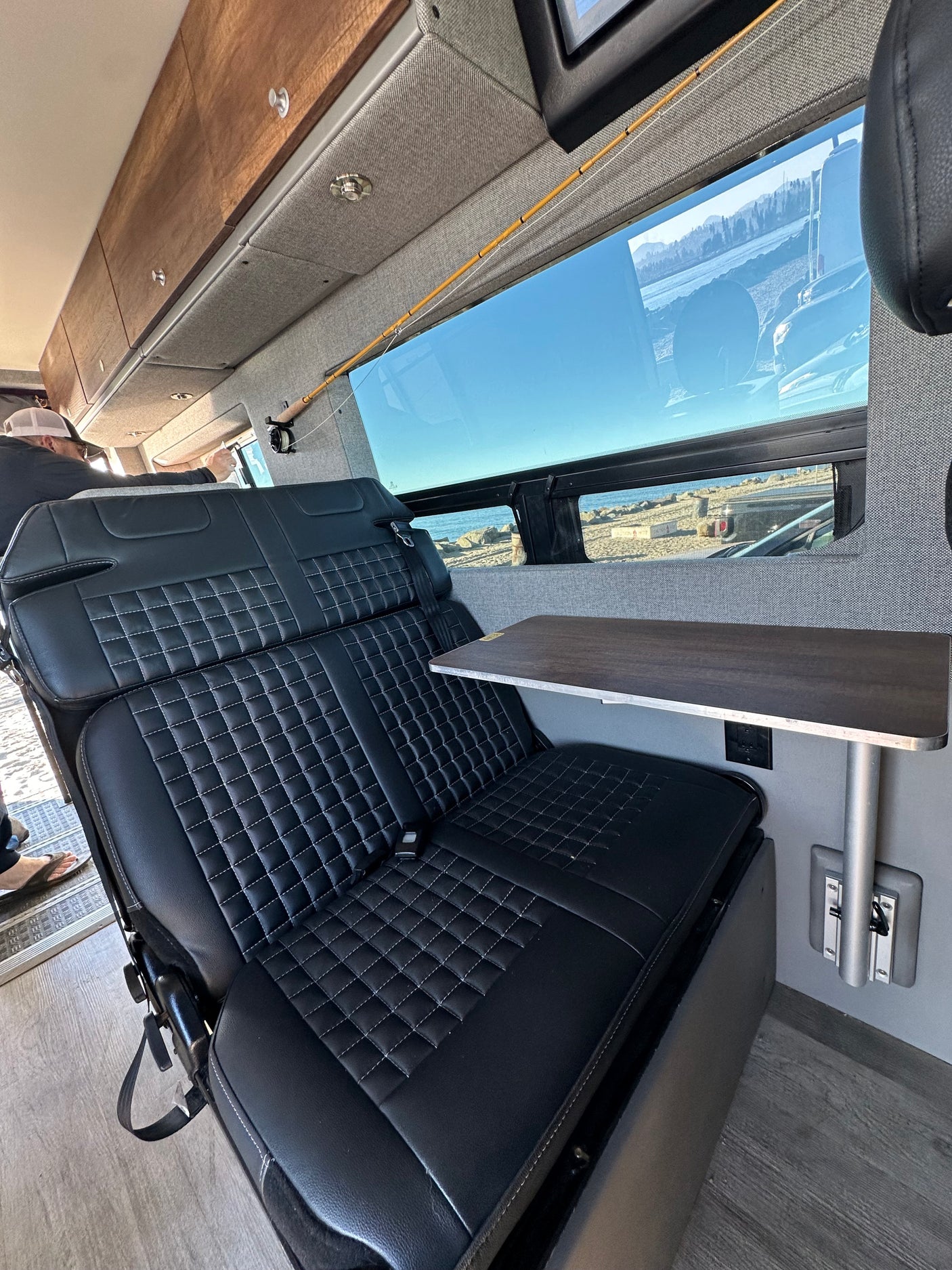 Spacious RV rental interior: black quilted seats, a side table, built-in fishing rod storage, and large windows revealing an ocean view.