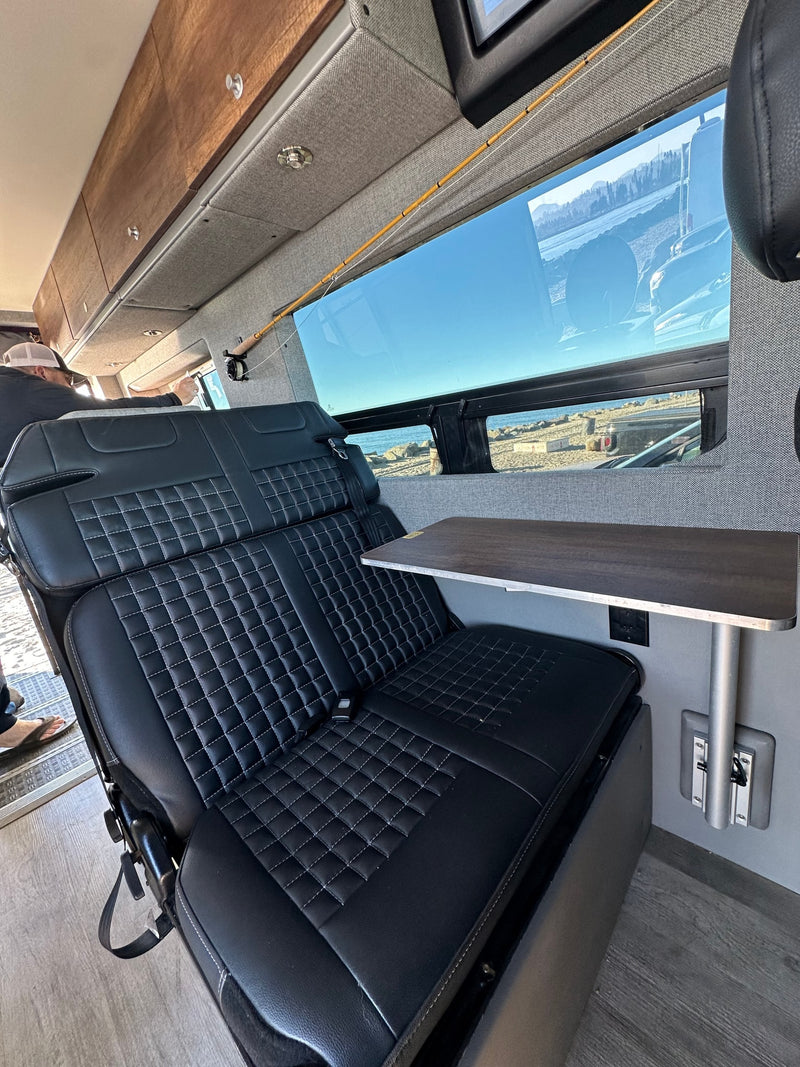 Spacious RV rental interior: black quilted seats, a side table, built-in fishing rod storage, and large windows revealing an ocean view. — photo 7