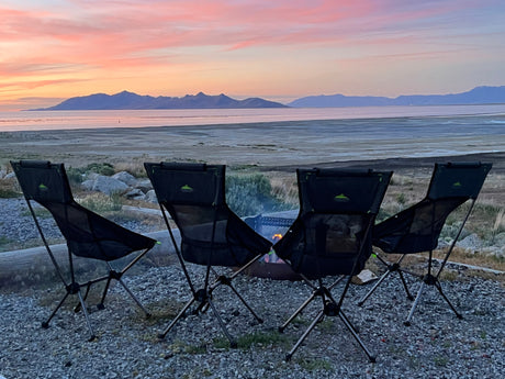 Four camp chairs by a fire pit, facing a beautiful sunset over distant mountains and a calm lake. Perfect RV rental site.