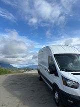 White RV rental van overlooks vast Alaskan landscape: mountains, river, and forests under cloudy sky.
