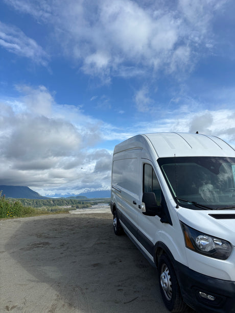 White RV rental van overlooks vast Alaskan landscape: mountains, river, and forests under cloudy sky.