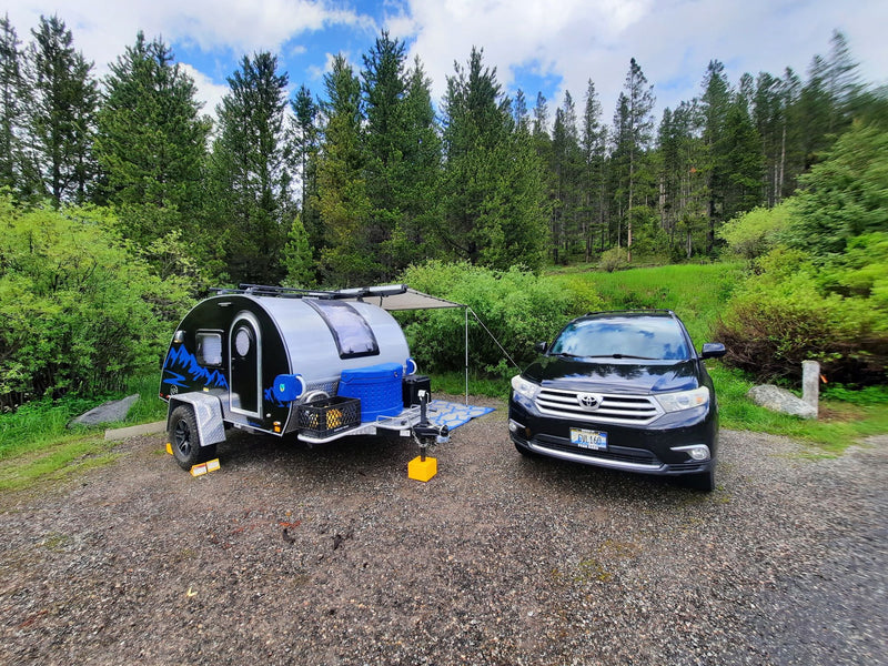 Black SUV & compact RV rental parked on a gravel site, framed by a lush green forest under a cloudy sky. — photo 1