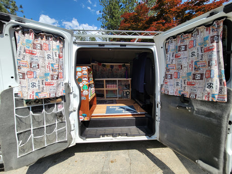 Cozy camper van interior, possibly an RV rental, featuring Route 66 curtains, custom shelving, and a "home sweet home" mat.