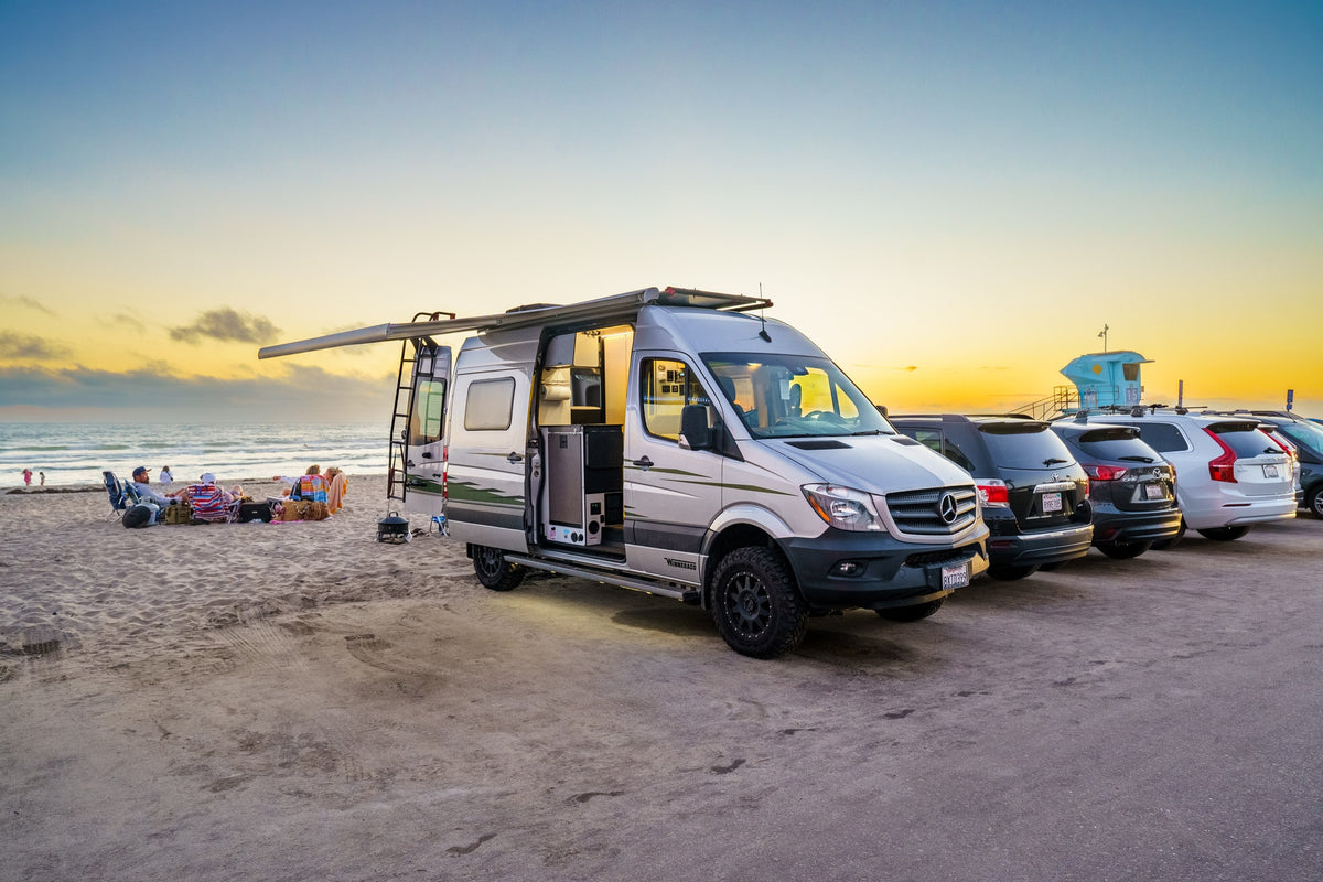 Relaxing beach sunset with an RV rental. A silver camper van is parked by the sand, awning extended, near people and the ocean.