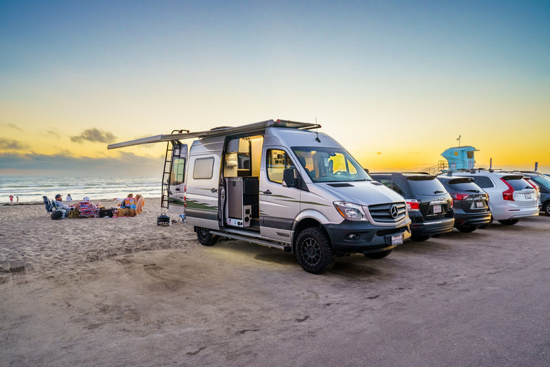 Relaxing beach sunset with an RV rental. A silver camper van is parked by the sand, awning extended, near people and the ocean. — photo 1