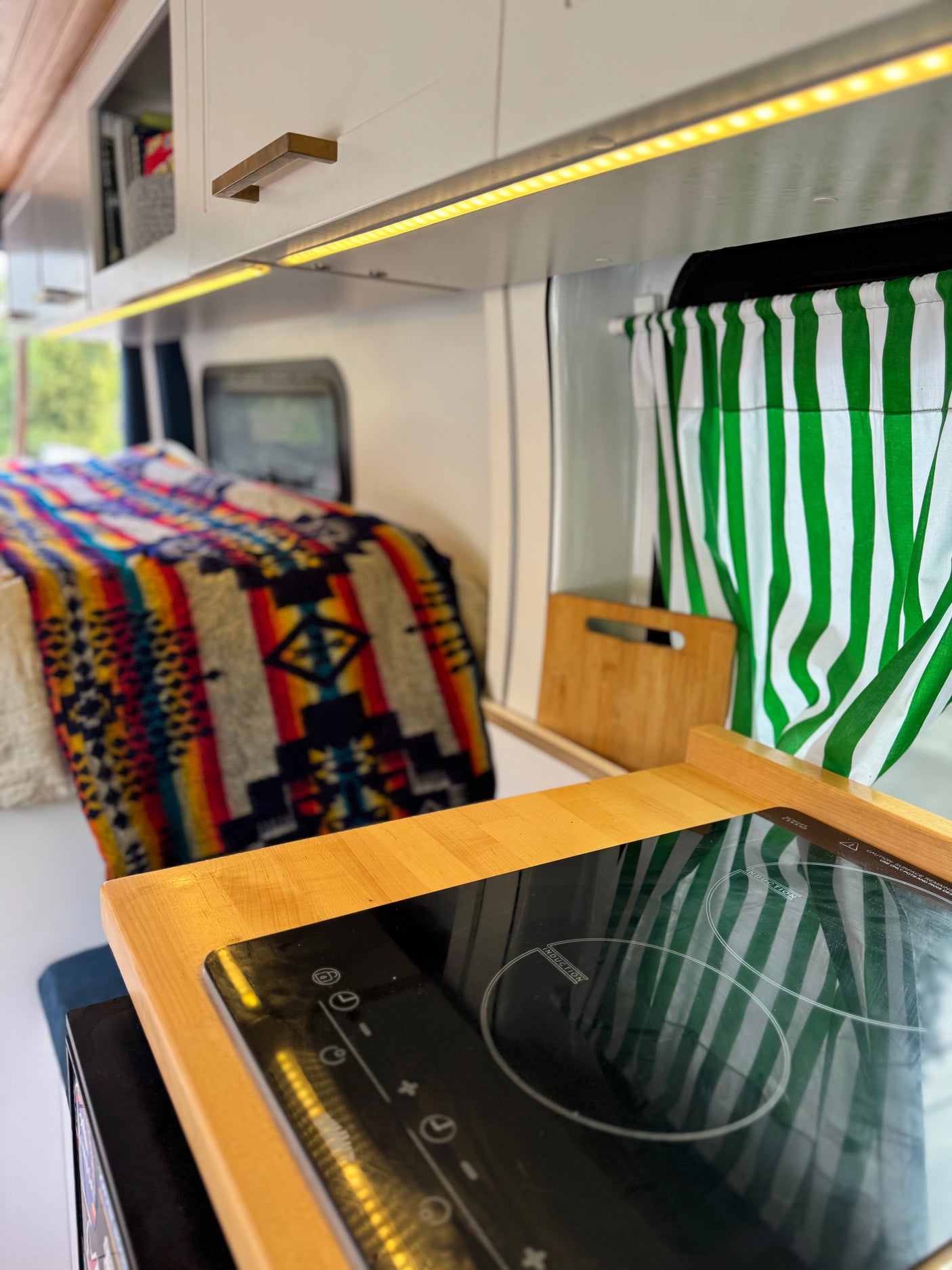 Cozy RV rental interior: Kitchenette with induction cooktop, bright lighting, green striped curtains, and a colorful bed.