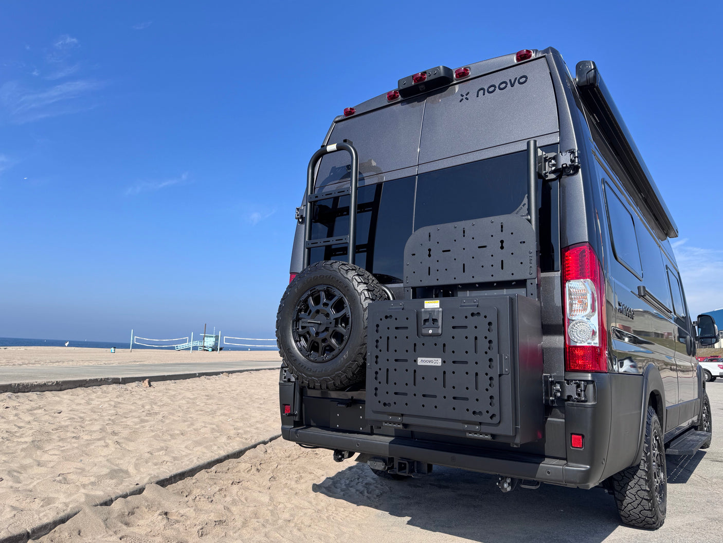 Dark grey Noovo X RV rental parked on a sandy beach. Rear features include a ladder, spare tire, and gear panels.