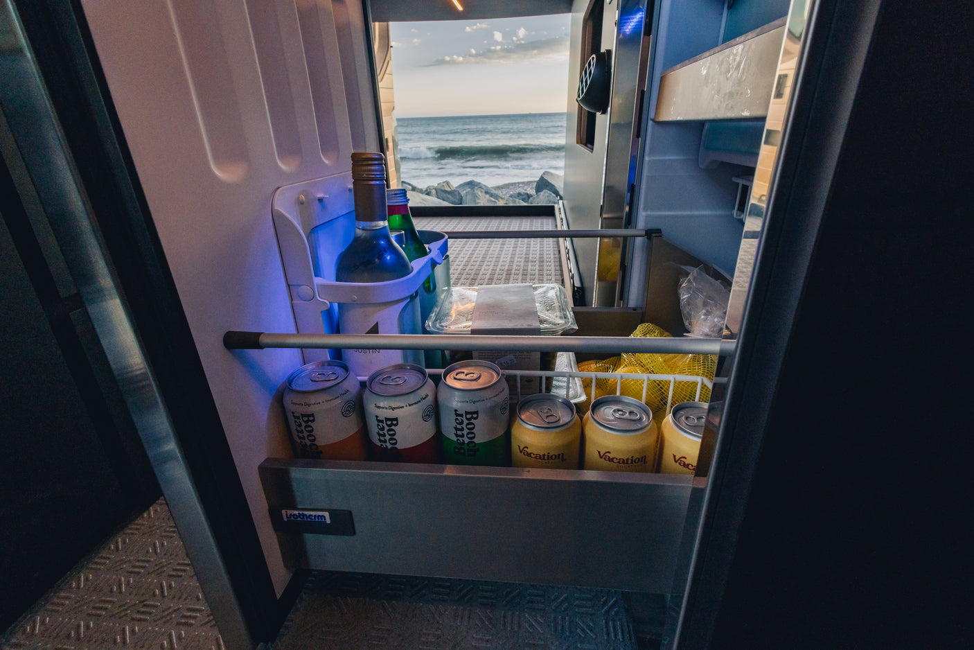 RV rental fridge stocked with beverages like kombucha & cocktails, open to a scenic ocean view.