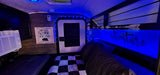 Cozy RV rental interior with vibrant blue LED lighting, bed, AC, TV, and rustic "Don't Wake the Bear" decor.