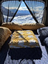 An RV rental's cozy interior, with a soft bed and pillows, offers breathtaking snowy mountain views.