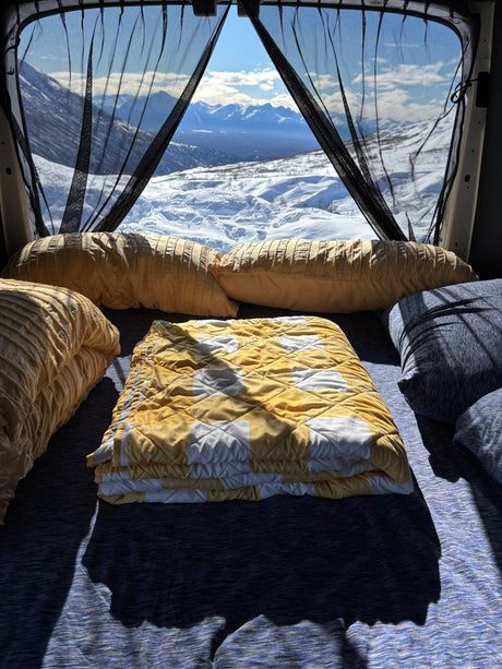 An RV rental's cozy interior, with a soft bed and pillows, offers breathtaking snowy mountain views.