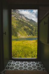 Escape with an RV rental! Gaze out the door at a vibrant mountain landscape with a serene lake, lush trees, and wildflowers.