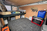 RV rental interior: cozy bed with under-bed storage, red portable stove, blue water jug, and a wooden bear decoration.