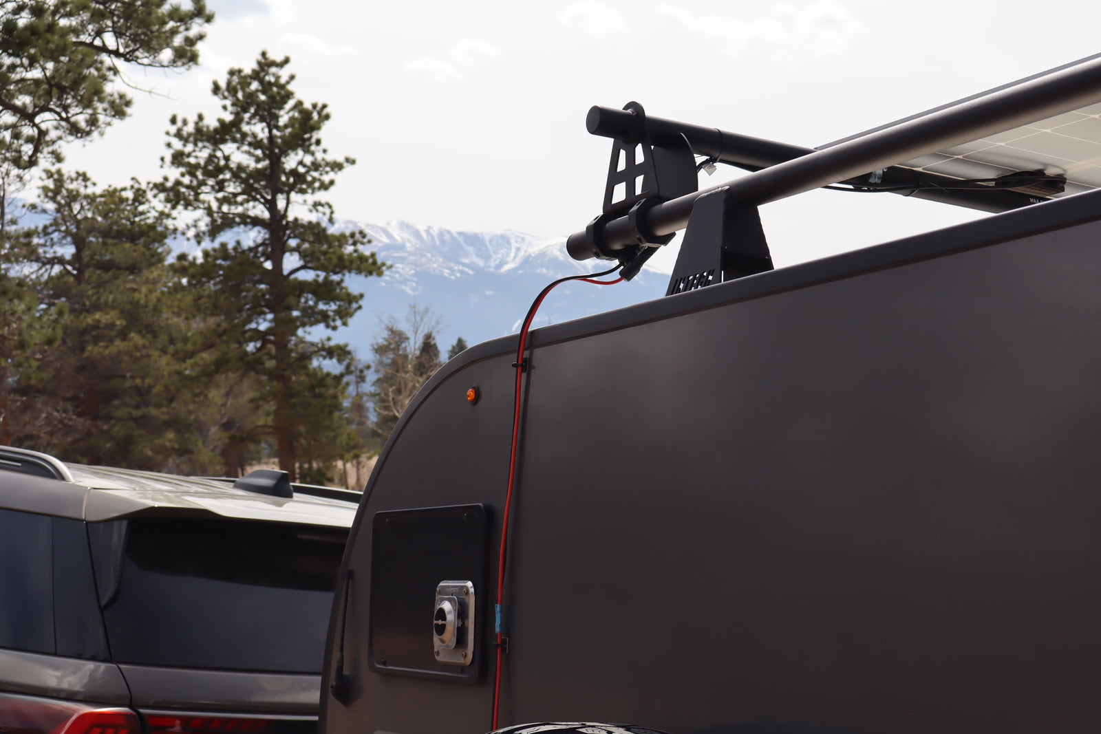 This rv rental shows a teardrop camper with solar panels, a roof rack, and an electrical hookup, set against scenic mountains.