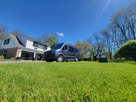 Modern dark gray Mercedes-Benz Sprinter van for rv rental, parked on a bright green lawn by a house.