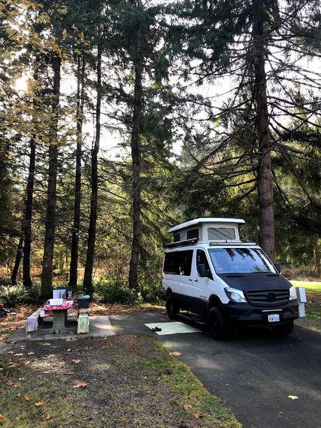 This white Mercedes Sprinter camper van, likely an rv rental, is set up at a peaceful forest campsite with a picnic table.