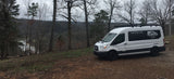 White Van Life KC RV rental on a dirt path overlooking a scenic wooded valley and lake.