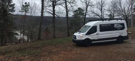 White Van Life KC RV rental on a dirt path overlooking a scenic wooded valley and lake.