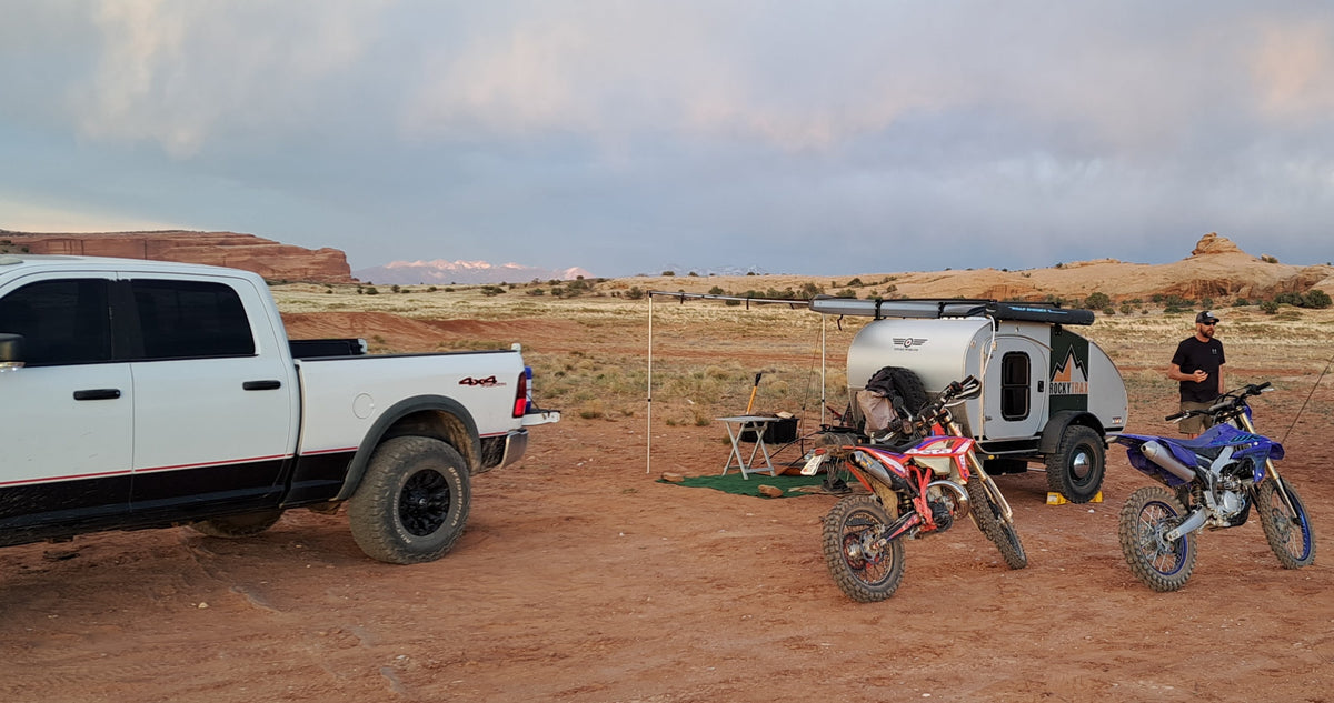 Desert RV rental adventure: truck, teardrop camper, & dirt bikes in a rugged landscape with red rocks & distant mountains. — photo 5