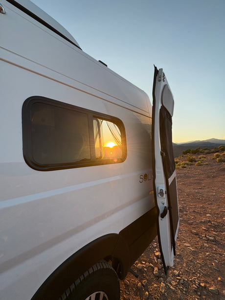 Sunset beams through the window of this white **RV rental** camper van, parked on rocky terrain with open rear doors.
