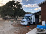 Silver Mercedes Sprinter RV rental leveled on a dirt lot, parked by a large oak tree and a barn.
