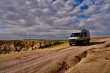 Modern RV rental van on a dirt road, overlooking badlands with communication towers under cloudy skies.