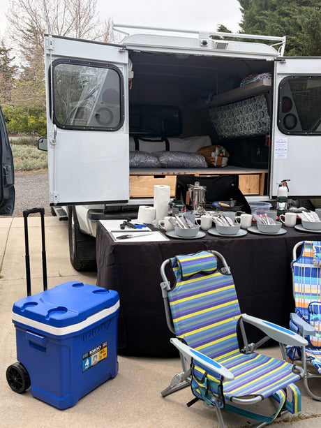 **RV rental** camper van with open rear doors, revealing bunk beds. An outdoor kitchen, cooler, and chairs are set up for camping.