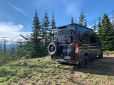 Scenic rv rental: A dark Ford Transit camper van is parked on a dirt path overlooking tall pine trees and mountain views.