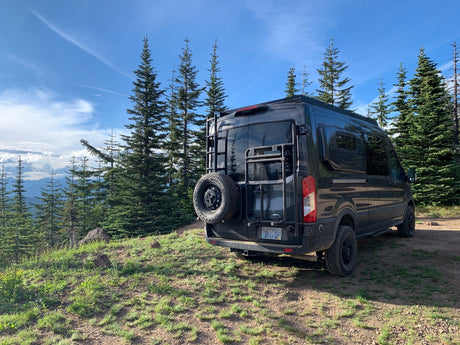Scenic rv rental: A dark Ford Transit camper van is parked on a dirt path overlooking tall pine trees and mountain views.