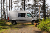 Silver Sprinter camper van parked among coastal trees at a 'Peace Vans Camp' site, perfect for RV rental adventures.