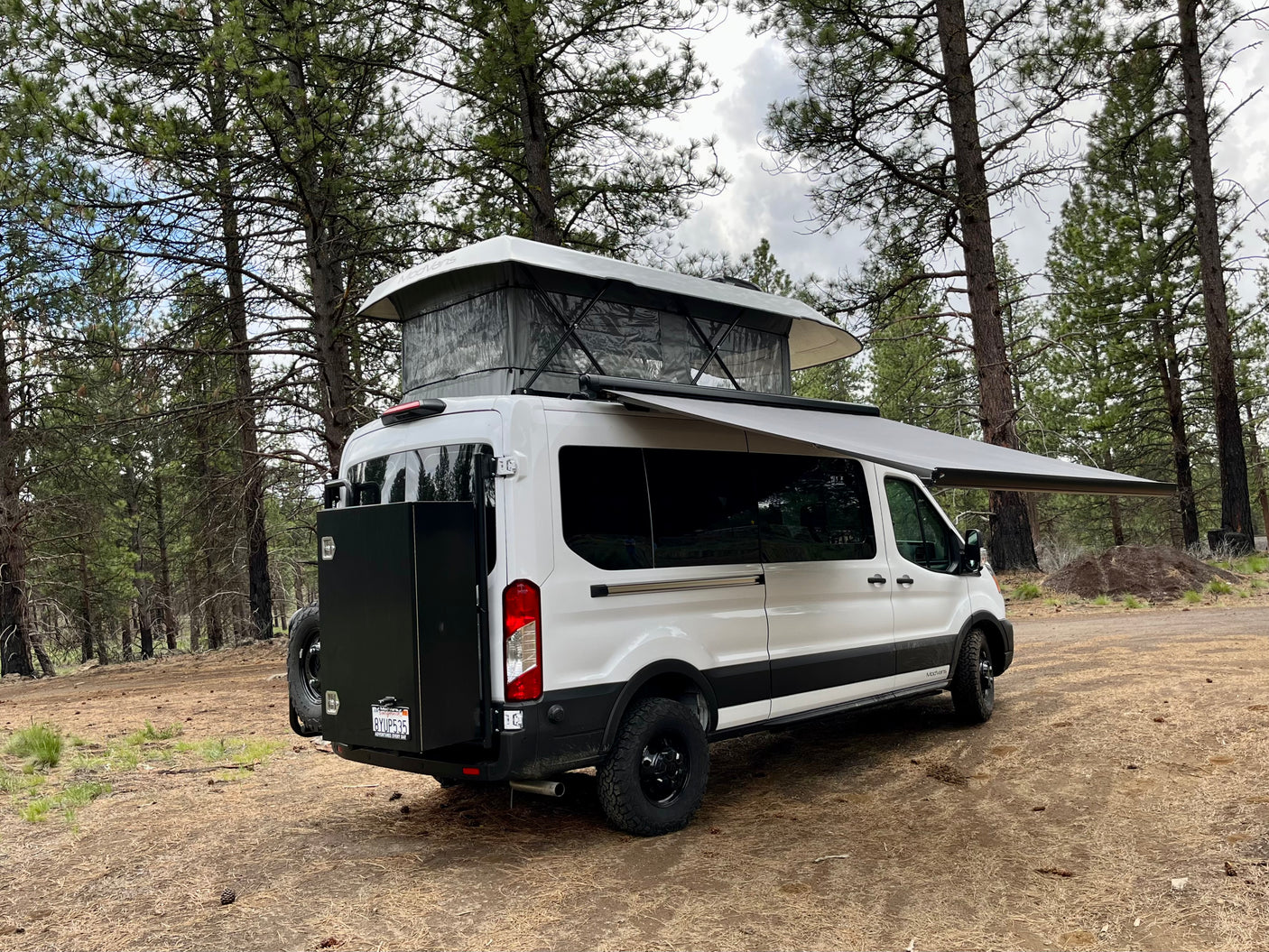 Potential RV rental: White camper van with pop-top roof & extended awning parked in a pine forest, ready for adventure.