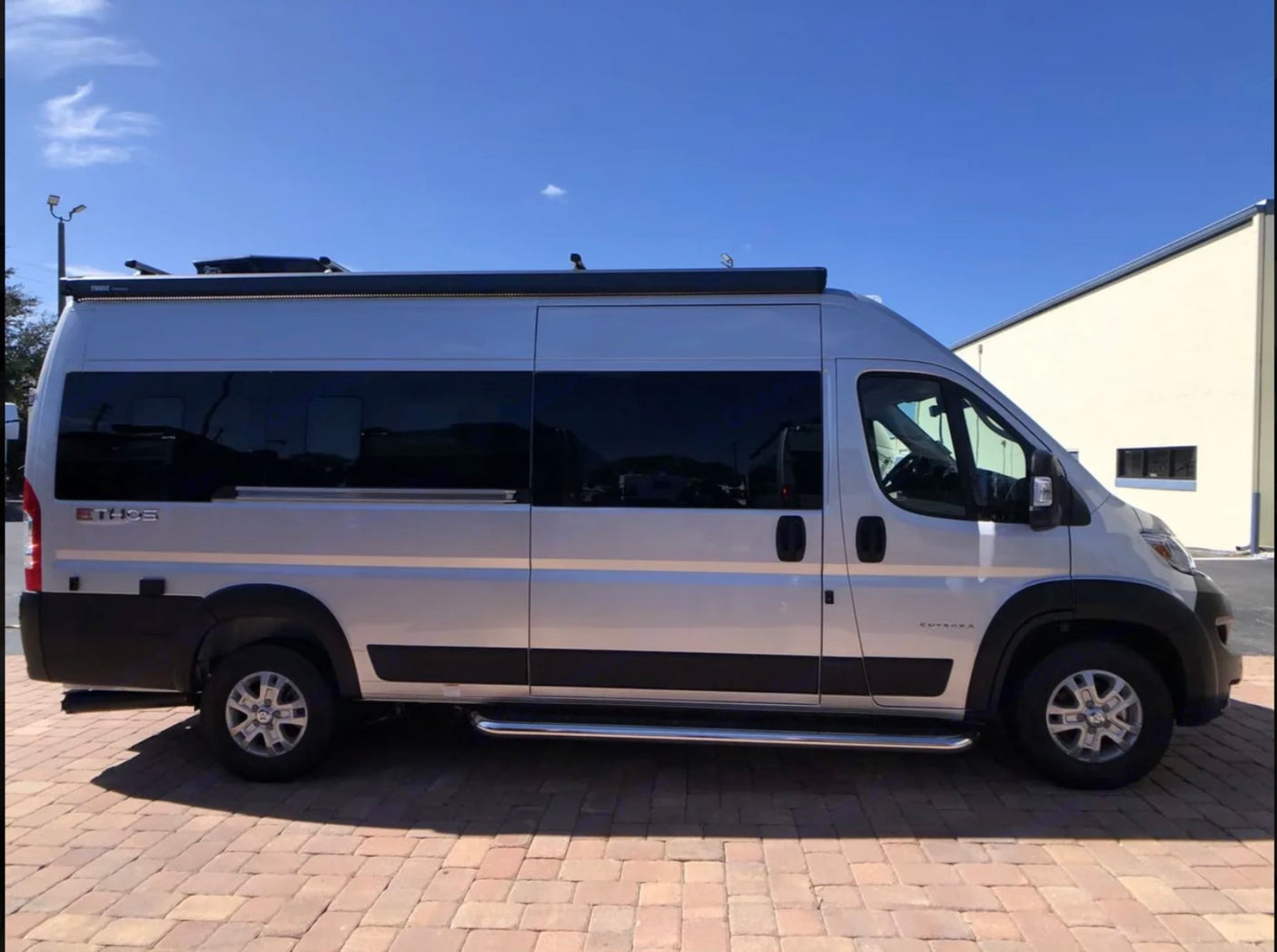 Side view of a silver camper van, ideal for RV rental, parked on pavers. Features include tinted windows and a roof awning.