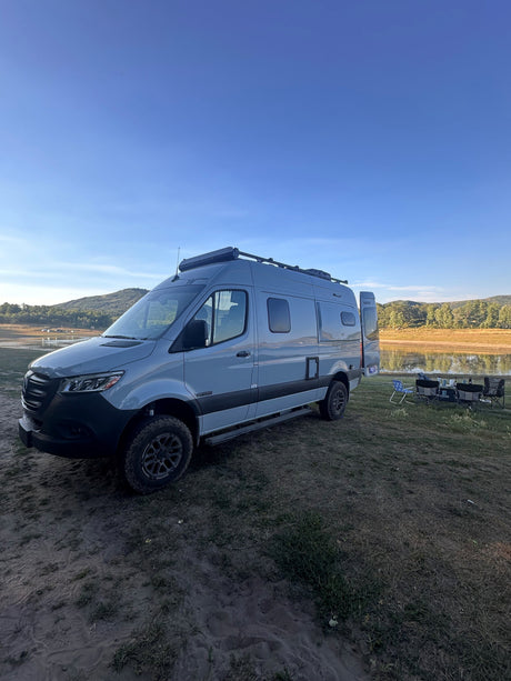 A grey RV rental is perfectly set up by a tranquil lake, with camping chairs and a table on the grassy bank.