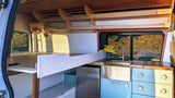 Inside a cozy RV rental: a modern kitchen with a brass sink, light blue cabinets, a comfortable bed, and custom wood shelving.