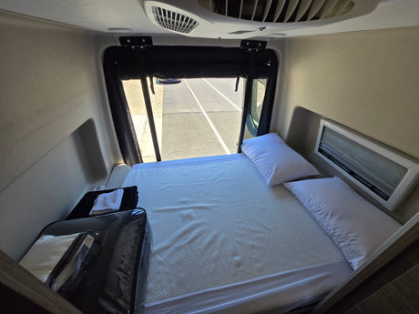 RV rental bedroom showcasing a bed with white pillows, a window view of the road, and an overhead AC unit.