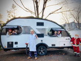 Joyful family holiday scene with an **RV rental**. Dad in PJs, mom & kids peeking from the festive teardrop camper windows, Santa nearby.