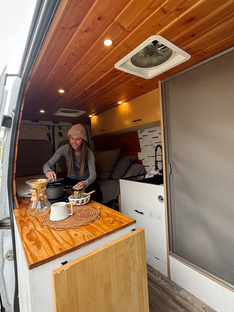 A woman smiles, preparing a meal & coffee in her cozy RV rental camper van. Perfect for van life adventures!