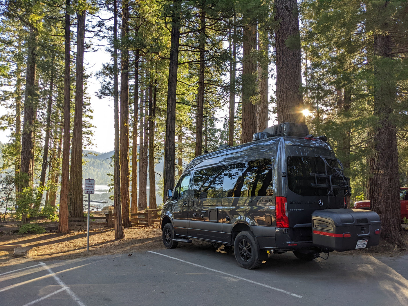 A gray rv rental camper van sits parked by a sunny lake and tall forest trees, hinting at outdoor adventures.